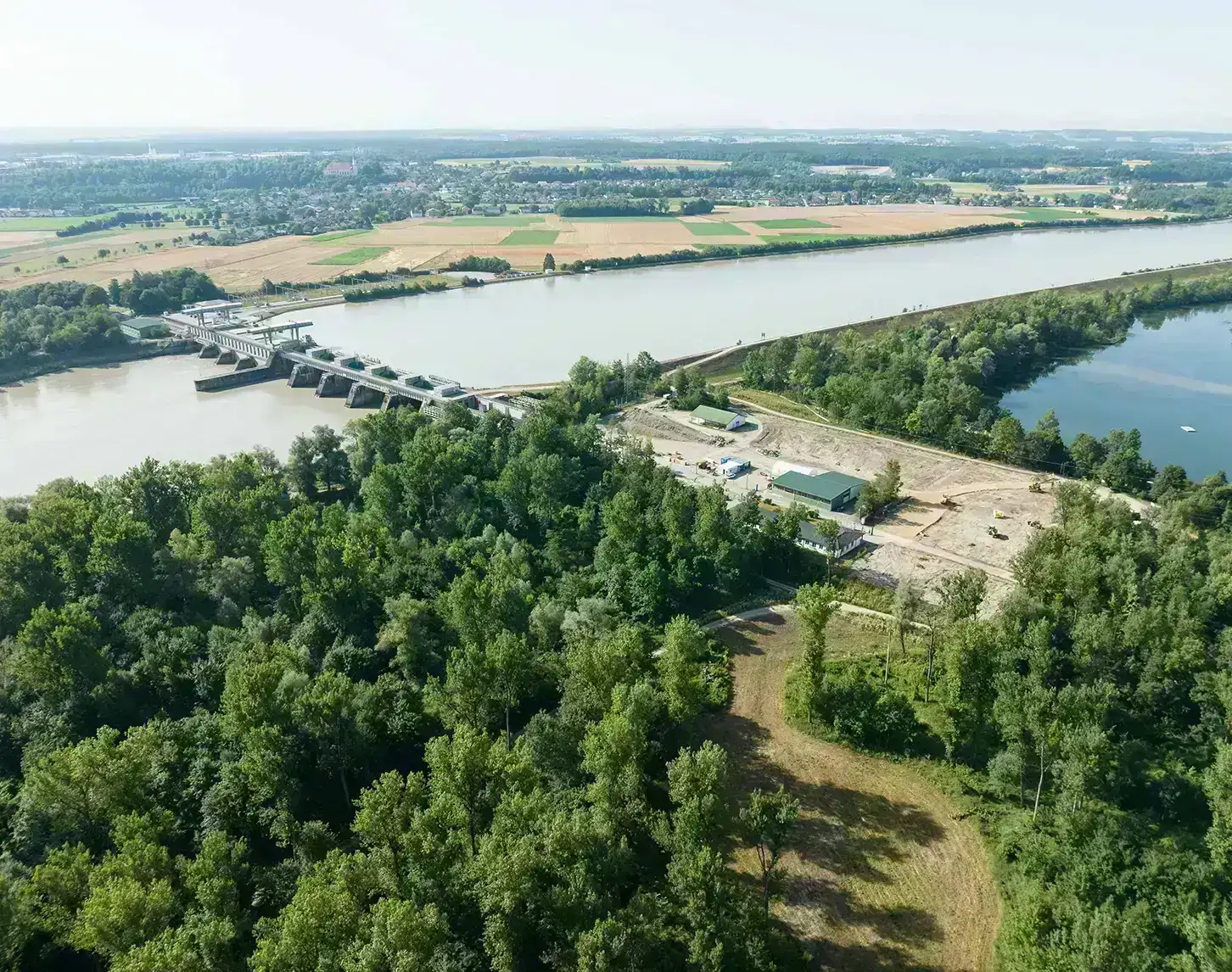 Aerial view of a river power plant, a forested area in the foreground and agricultural fields in the background.